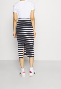 Kvinna klädd i en vit t-shirt, en marinblå och vit horisontell randig midi-kjol med en slits baktill, vita strumpor och vita sneakers med röda klackar.