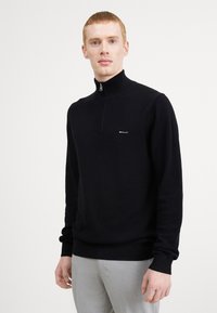 Zwarte zip-nekkpullover van textuur gebreide stof, met een klein logo op de borst en ribgebreide manchetten en onderkant. Model draagt lichte grijze broek.