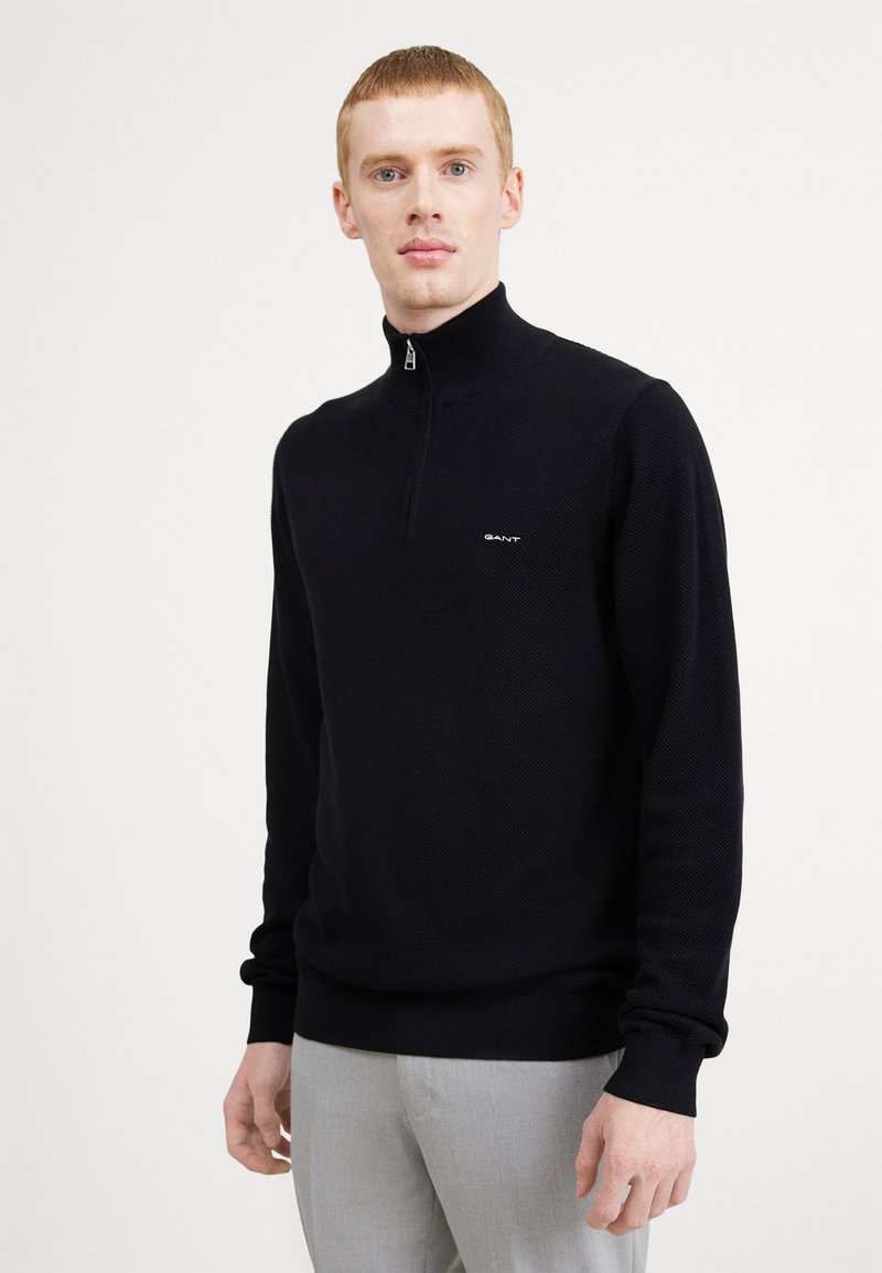 Zwarte zip-nekkpullover van textuur gebreide stof, met een klein logo op de borst en ribgebreide manchetten en onderkant. Model draagt lichte grijze broek.