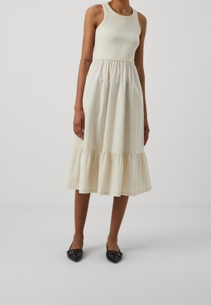 Kobieta w kremowej, bezrękawnej sukience midi z marszczonym dołem i falbaną na krawędzi, w zestawieniu z czarnymi balerinami w czubek.