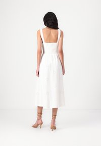 Vista posteriore di un vestito bianco senza maniche con un corpetto aderente, vita arricciata e disegni di sole ricamati. Completato da sandali con cinturini.