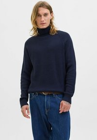 Navy ribbed turtleneck sweater with long sleeves, featuring a relaxed fit. Paired with blue denim trousers and a leather belt.
