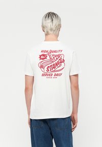White cotton t-shirt features a red graphic on the back with the text “HIGH QUALITY No.1 STANCE SERVED DAILY SINCE 2009” and a hotdog illustration.