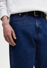 Jeans de mezclilla azul oscuro con corte recto, costuras visibles y bolsillos delanteros. Una mano descansa al lado de la cinturilla, destacando un cinturón negro.