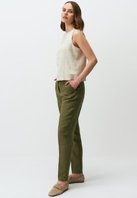 Helles beige ärmelloses Oberteil kombiniert mit olivgrünen, taillierten Hosen. Der Stoff wirkt weich mit einer dezenten Textur. Legere Slipper in Beige.