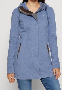 Felpa blu con zip, cappuccio con cordini e dettagli marroni sul colletto. Presenta due tasche laterali con zip.