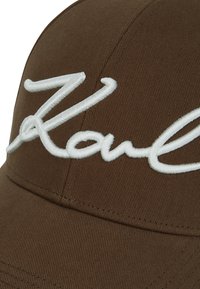 Καφέ ύφασμα καπέλο μπέιζμπολ με μεγάλο λευκό κεντημένο λογότυπο σε καλλιγραφία στην μπροστινή πάνελ.