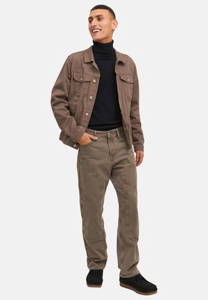 Hymyilevä mies, jolla on päällään ruskea napillinen takki, musta pooloneule, ruskeat housut paikkataskuilla ja mustat kengät, seisomassa käsi taskussa.