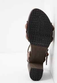 Sandale en cuir marron avec une semelle en caoutchouc noir texturé, arborant un design courbé et des accents ronds décoratifs sur les sangles.