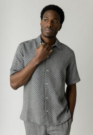 Mand iført kortærmet skjorte med knapper og matchende bukser med lille geometrisk mønster, poserer med den ene hånd nær kraven mod en ensfarvet baggrund.