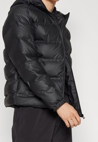 Černá péřová bunda se zipem, kapucí a pružnými manžetami. Materiál má hladký, lesklý povrch s prošitým vzorem.