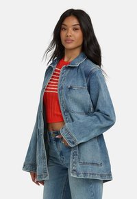 Veste en denim de couleur bleu clair avec une coupe décontractée, dotée de deux poches avant et d'une fermeture à boutons. Associée à un crop top rouge à côtes.