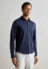 Camicia button-up a maniche lunghe di colore navy, in tessuto morbido, colletto classico, texture liscia, leggera lucentezza e chiusura frontale con bottoni, abbinata a pantaloni chiari.