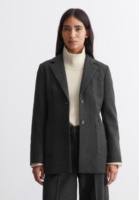 Kivikharja hall kašmiri koos klapiga krae, ühekohaline nööp kinnitusega ja kahe esiküljel oleva taskuga. Tekstureeritud villane kangas, kantud kreemjas turtlenecki üle.