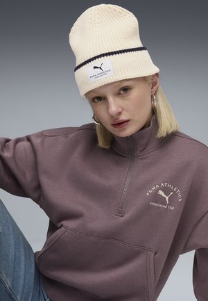 Purple zip-up sweatshirt with logo, and a ribbed cream beanie with navy stripe. Model wears hoop earrings. Denim visible at the bottom.