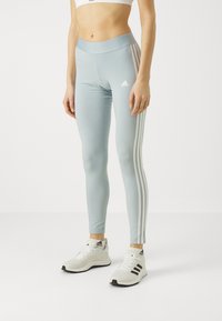 Ljusblå leggings med hög midja, som har tre vertikala vita ränder längs varje sida. Kombineras med vita sportskor.