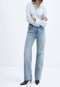 Persona con camisa de botones azul claro, jeans de tiro alto azul claro, botas de tacón puntiagudas negras, de pie con los brazos cruzados.