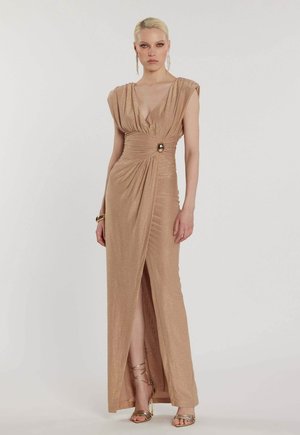 Donna con capelli biondi che indossa un lungo abito beige scintillante con scollatura profonda a V, spacco laterale, dettaglio in vita, orecchini dorati, braccialetto e tacchi con cinturini.