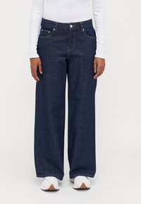 Mörkblå vida jeans i denim, med främre fickor, knappstängning och synliga sömmar. Stylade med vita sneakers.