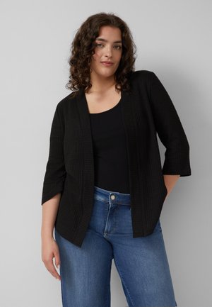 Frau mit lockigem Haar, die eine schwarze, strukturierte offene Strickjacke über einem schwarzen Oberteil und blauen Jeans trägt, steht vor einem schlichten grauen Hintergrund.