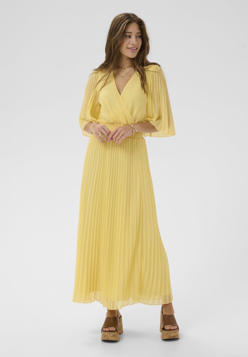 Femme portant une longue robe jaune plissée avec des manches amples et des sandales compensées marron, debout devant un fond gris uni.