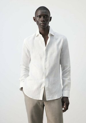 Homme portant une chemise blanche à manches longues boutonnée et un pantalon beige clair, se tenant devant un fond uni clair.