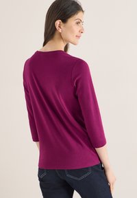 Chemise magenta à manches trois-quarts avec un col rond, une texture lisse et une coupe décontractée, associée à un jean foncé avec des coutures contrastantes.