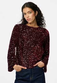 Top lungo a maniche con paillettes bordeaux, dalla morbida texture in velluto, scollatura rotonda e vestibilità comoda, abbinato a jeans in denim scuro.