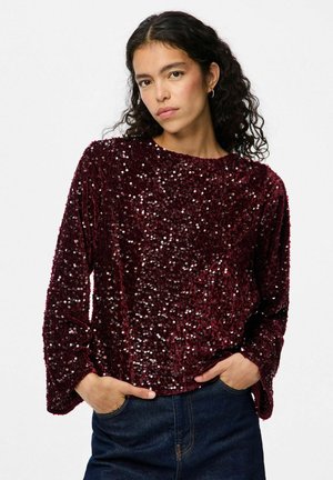 Top lungo a maniche con paillettes bordeaux, dalla morbida texture in velluto, scollatura rotonda e vestibilità comoda, abbinato a jeans in denim scuro.