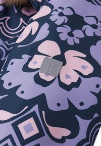 Patrón floral en púrpura y rosa sobre un fondo azul oscuro, con un parche cuadrado texturizado que presenta un logo en relieve de "R" en la esquina.