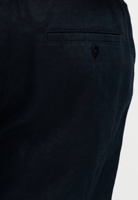 Gros plan sur un pantalon en tissu sombre montrant une poche arrière avec un seul bouton noir et une couture horizontale.