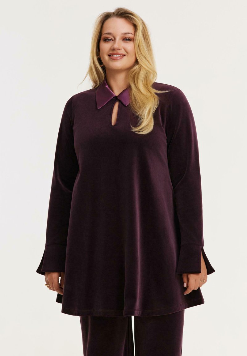 Glunderende vrouw met lang blond haar, gekleed in een donkerpaarse fluwelen tuniek met lange mouwen en een keyhole-halslijn, met bijpassende broek.