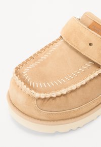 Chaussure en daim beige avec une forme arrondie, des détails cousus et une sangle à fermeture auto-agrippante. Dispose d'une semelle texturée pour une bonne adhérence.