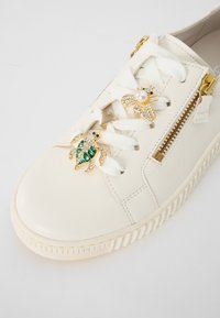 Zapatilla de cuero blanco con acentos decorativos de oro en forma de abeja y tortuga, cordones blancos y una cremallera lateral. Detalle de suela texturizada visible.