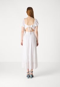 Vit klänning med puffärmar, öppen rygg knuten med rosetter, rynkad midja och flerstegs fåll. Matchas med ljusblå sandaletter med klack.