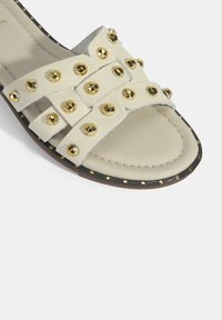 Slide-sandaal van crèmekleurig leer, voorzien van gouden studs, een gevlochten bandontwerp, een platte zool en een gestructureerde voetbed.