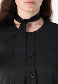 Black blouse with a smooth texture, featuring a tie detail at the neck, round neckline, and black buttons down the front.