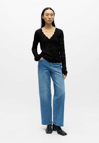 Schwarze gestrickte Strickjacke mit gewellten Kanten und Knopfverschluss, kombiniert mit hellblauen, weit geschnittenen Jeans und schwarzen, eckigen Ankle Boots.