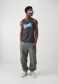 Svart tank topp med "Hilsener fra Levi's" grafikk, grå cargo-bukser og lavendel-sneakers. Store hvite solbriller fullfører antrekket.