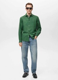 Groene linnen button-up shirt met een borstzak, gecombineerd met lichtblauwe denim jeans en zwarte schoenen. Het model draagt zwarte zonnebril.