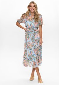 Mujer sonriendo con cabello rubio suelto, vistiendo un vestido midi abotonado de flores pastel con mangas cortas y cinturón en la cintura, combinado con sandalias amarillas.