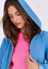 Niebieska bluza z zamkiem błyskawicznym, z podszytym kapturem i kontrastującym niebieskim wykończeniem, noszona na różowym swetrze o fakturowej strukturze. Miękka tkanina i luźny krój.