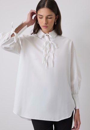 White blouse with a high collar, tied with thin white ribbons at the neckline and sleeves. Features a loose fit and smooth texture.