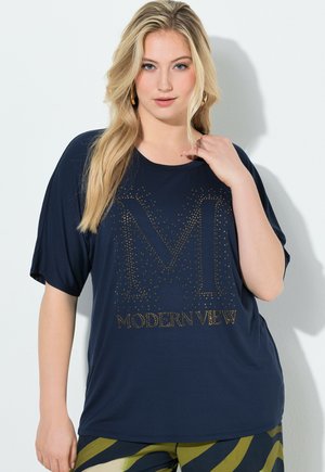 Femme blonde portant un t-shirt bleu marine avec le texte "M MODERN VIEW" en strass dorés et un pantalon à motifs verts, debout contre un mur blanc.