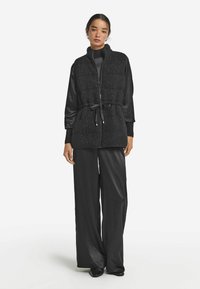 Quiltad svart väst med dragkedja och justerbart knytmidja. Kombinerad med en elegant svart satinblus och vida byxor. Slät textur överlag.