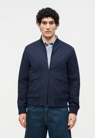 Man with curly hair wearing a navy zip-up jacket over a light blue button-up shirt and blue jeans, standing against a plain background.