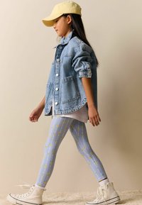 Džínová bunda s velkými kapsami a knoflíky, světle modré legíny se žlutými květinovými vzory a bílé vysoké tenisky. Na hlavě žlutá čepice.