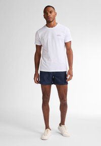 Tricou alb cu mânecă scurtă, cu text mic roșu; asociat cu pantaloni scurți bleumarin; model purtând adidași albi; fundal simplu.