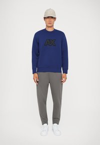 Felpa blu con logo nero, pantaloni grigi e sneakers bianche.Il cappellino è di un beige chiaro con un logo sul davanti. Stile minimalista.
