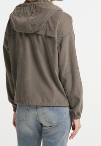 Khaki letvægtsjakke med snoretræk i hætten, elastiske manchetter og en afslappet pasform; båret over blå denim jeans. Blød tekstur, minimalistisk design.